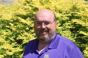 Mr. Hatch smiling in front of a shrub and wearing a purple Broughton polo shirt